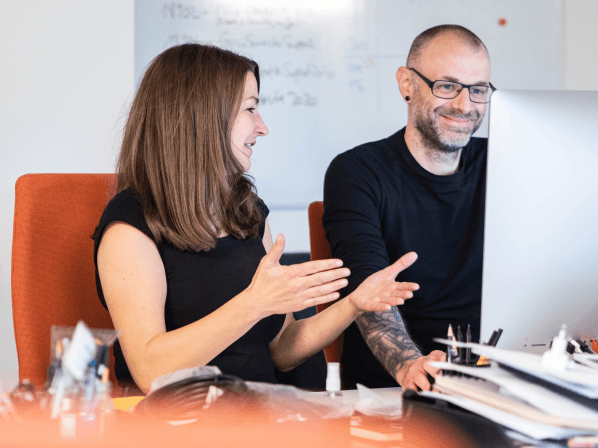 Andrea und Robert vorm Monitor. Andrea erklärt und zeigt etwas mit den Händen. 