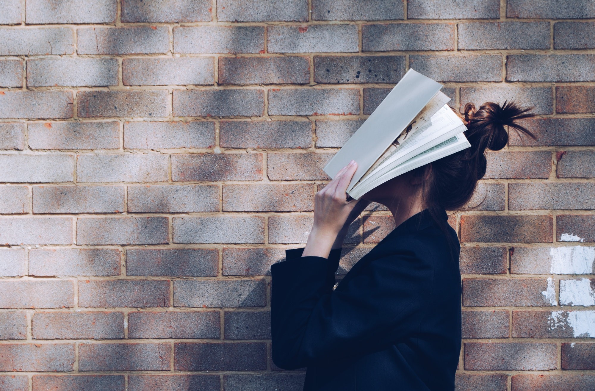 Frau steht vor einer Steinmauer und hält sich ein Buch vor das Gesicht. 