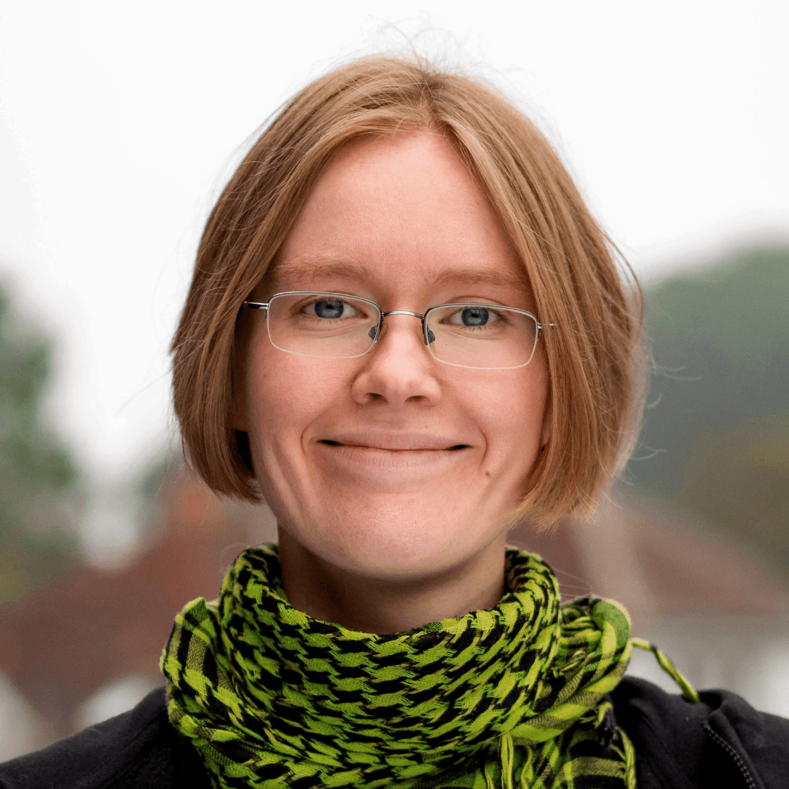 Anja hat kinnlanges blondes Haar und trägt eine Brille mit weißem Gestell und einen grünen Schal.