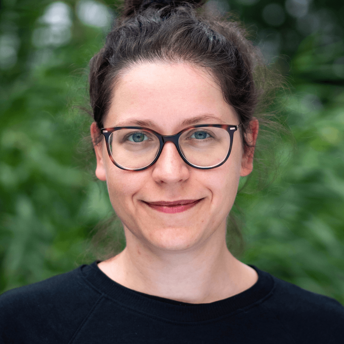 Annas dunkle Haare sind zusammengebunden und sie trägt eine Brille mit großen Gläsern und dunklem Gestell.