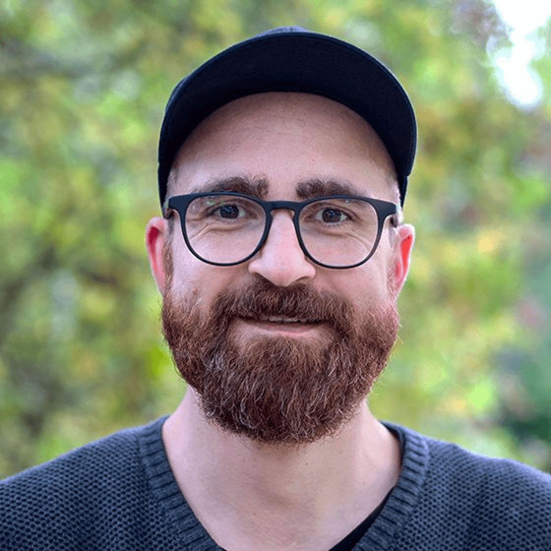 Christian hat einen Vollbart, trägt ein Cappy und eine Brille mit großen rundlichen Gläsern und schwarzem Gestell.