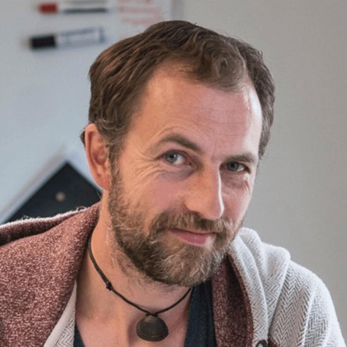 Enrico hat kurzes Haar und einen Vollbart und zieht leicht lächelnd eine Augenbraue hoch.