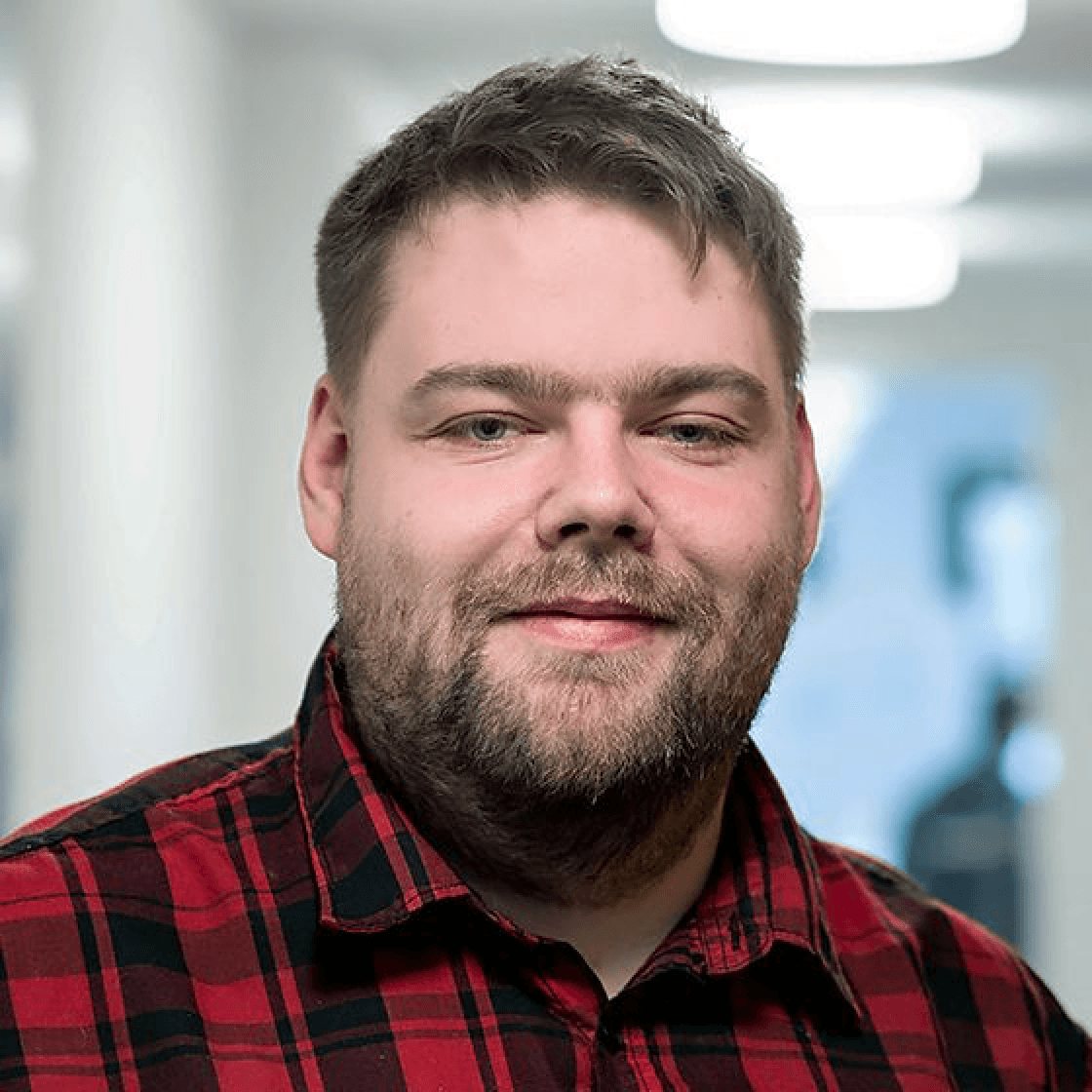 Florian hat kurzes blondes Haar, einen Vollbart und trägt ein rot-schwarz-kariertes Hemd. Er lächelt ganz leicht.