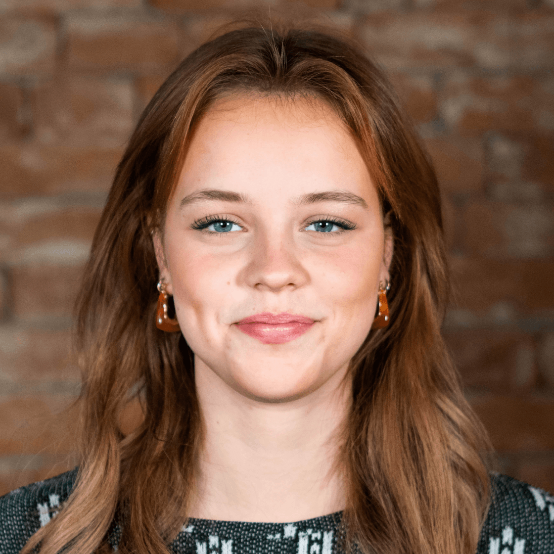 Greta hat langes braunes Haar, das ihr auf die Schultern fällt. Sie lächelt leicht.