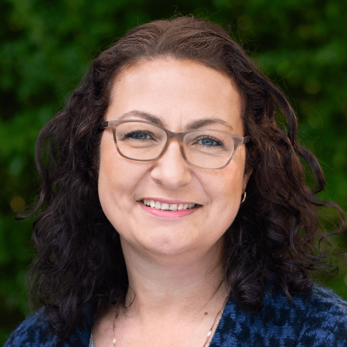 Ina hat lockiges dunkles Haar, trägt eine Brille und lächelt freundlich.