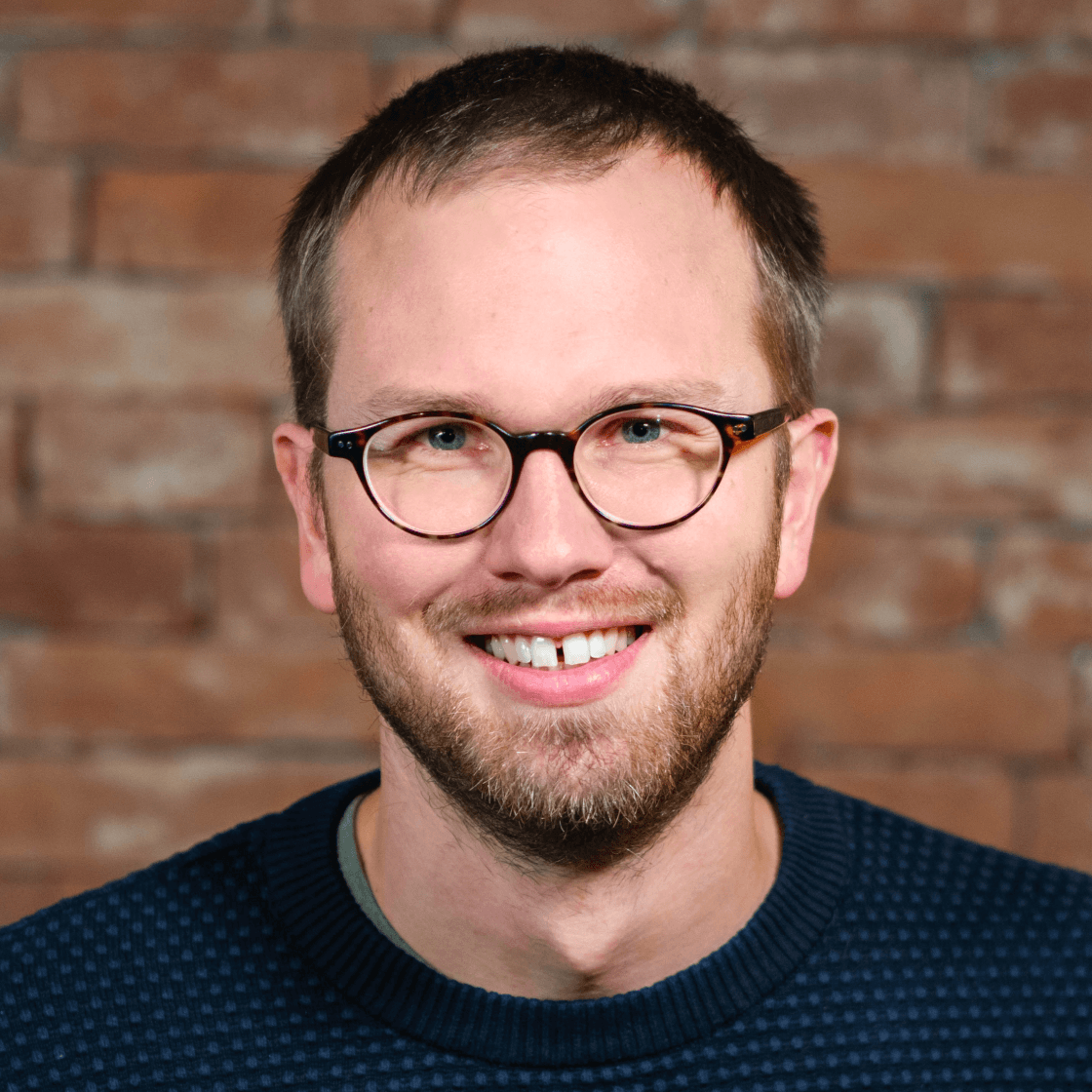 Jan hat kurzes dunkles Haar und eine Brille mit schwarzem Gestell. Er lächelt freundlich und hat eine kleine Lücke zwischen den oberen Schneidezähnen.
