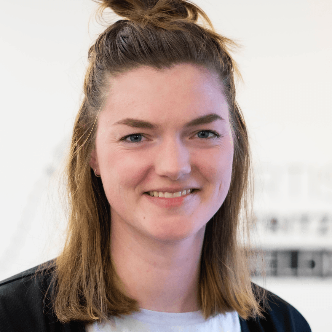 Melina hat braunes Haar, von denen die oberen Strähnen zusammengebunden sind und lächelt leicht.
