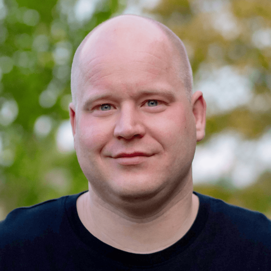 Paul trägt eine Glatze, ein schwarzes Shirt und lächelt leicht in die Kamera. 
