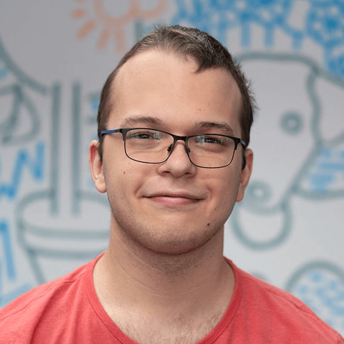 Ruchard hat kurze, dunkelblonde Haare, eine Brille und lächelt in die Kamera. Er trägt ein rotes Shirt. 