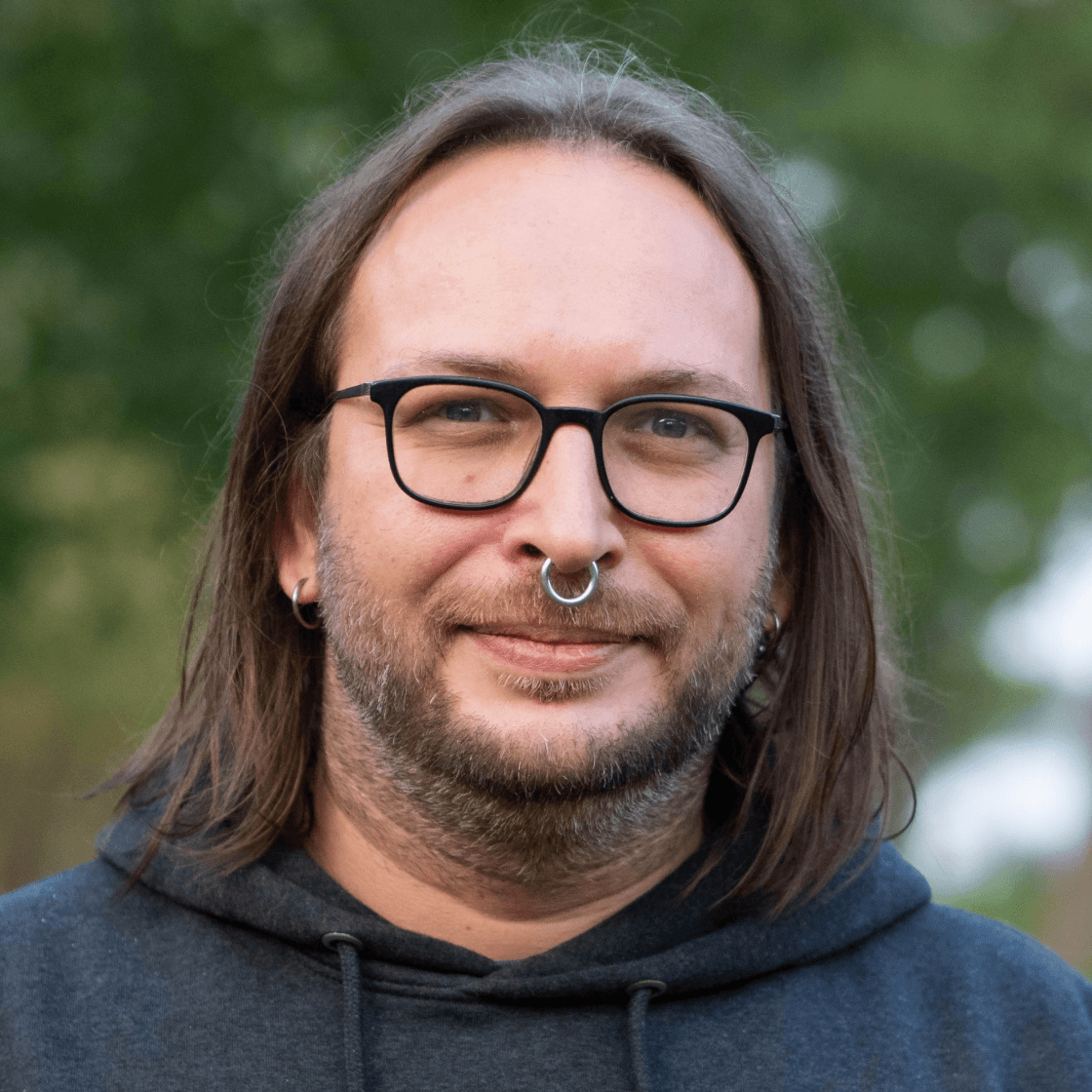 Robert trägt braune, schulterlange Haare, silberne Ohrringe und einen großen Nasenring. Er lächelt durch seinen Dreitagebart und hat eine große, schwarze Brille auf der Nase. 