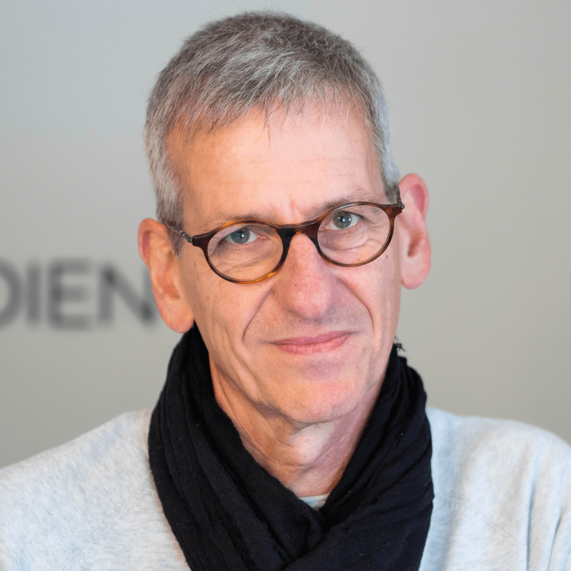 Rüdiger hat kurze, hellgraue Haare und eine runde, braune Brille. Er lächelt leicht in die Kamera und trägt einen schwarzen Schal und hellgrauen Pulli. 