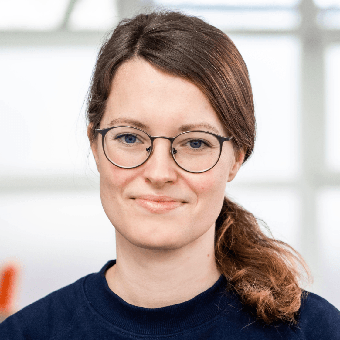 Susi trägt ihre dunkelblonden Haare zu einem Zopf gebunden. Sie hat eine runde Brille, blau-graue Augen und lächelt leicht in die Kamera. 