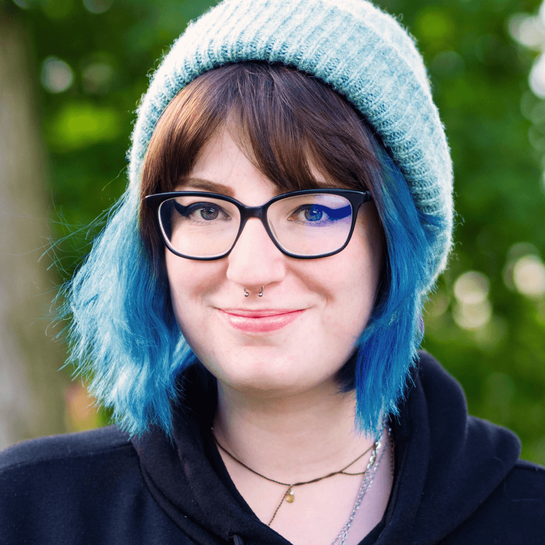 Valeria trägt eine blaue Mütze und passend dazu blaue gesträhnte Haare. Sie hat eine große, schwarze Brille und lächelt in die Kamera.