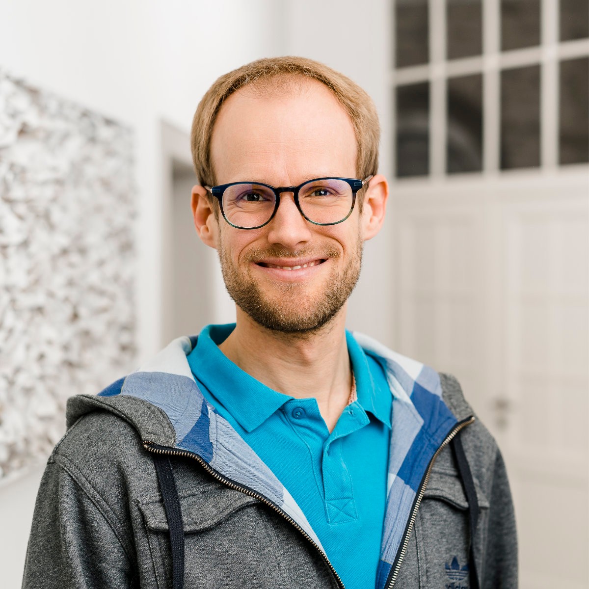Daniel strahlt mit einem breiten Lächeln in die Kamera. Er trägt kurzes, mittelblondes Haar, eine dunkle Brille und einen Dreitagebart. 