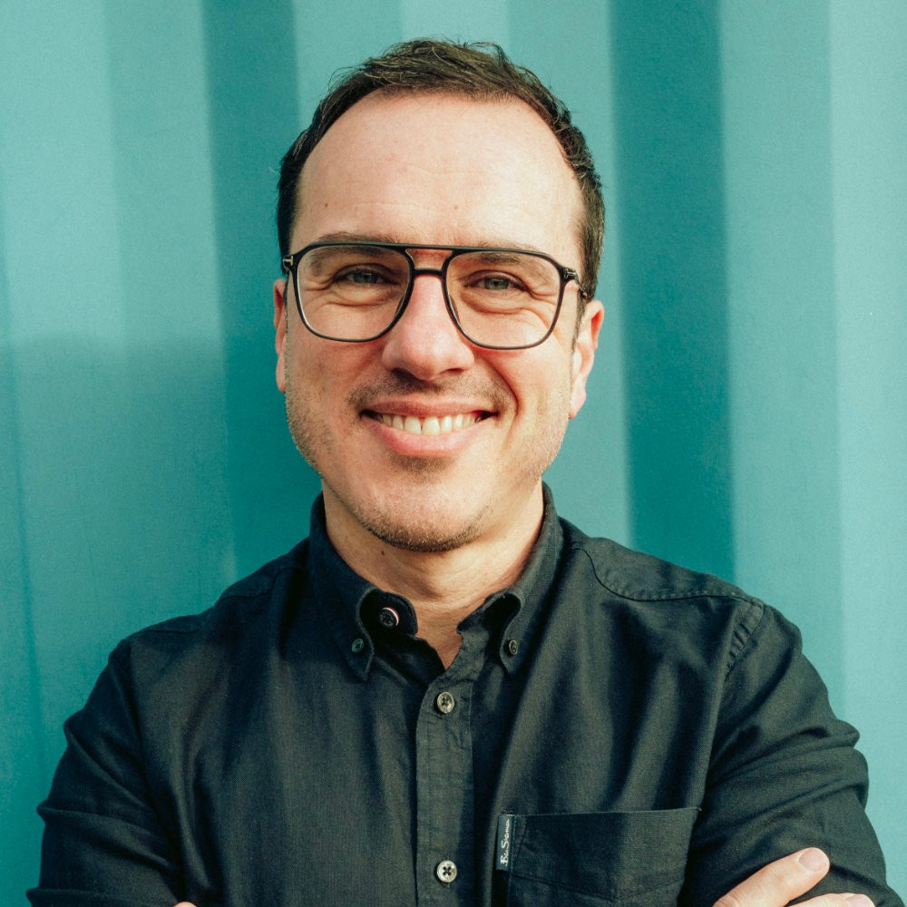 Daniel Hoffmann trägt eine dunkle, große Brille, kurzes braunes Haar, trägt ein schwarzes Hemd und lächelt in die Kamera. 