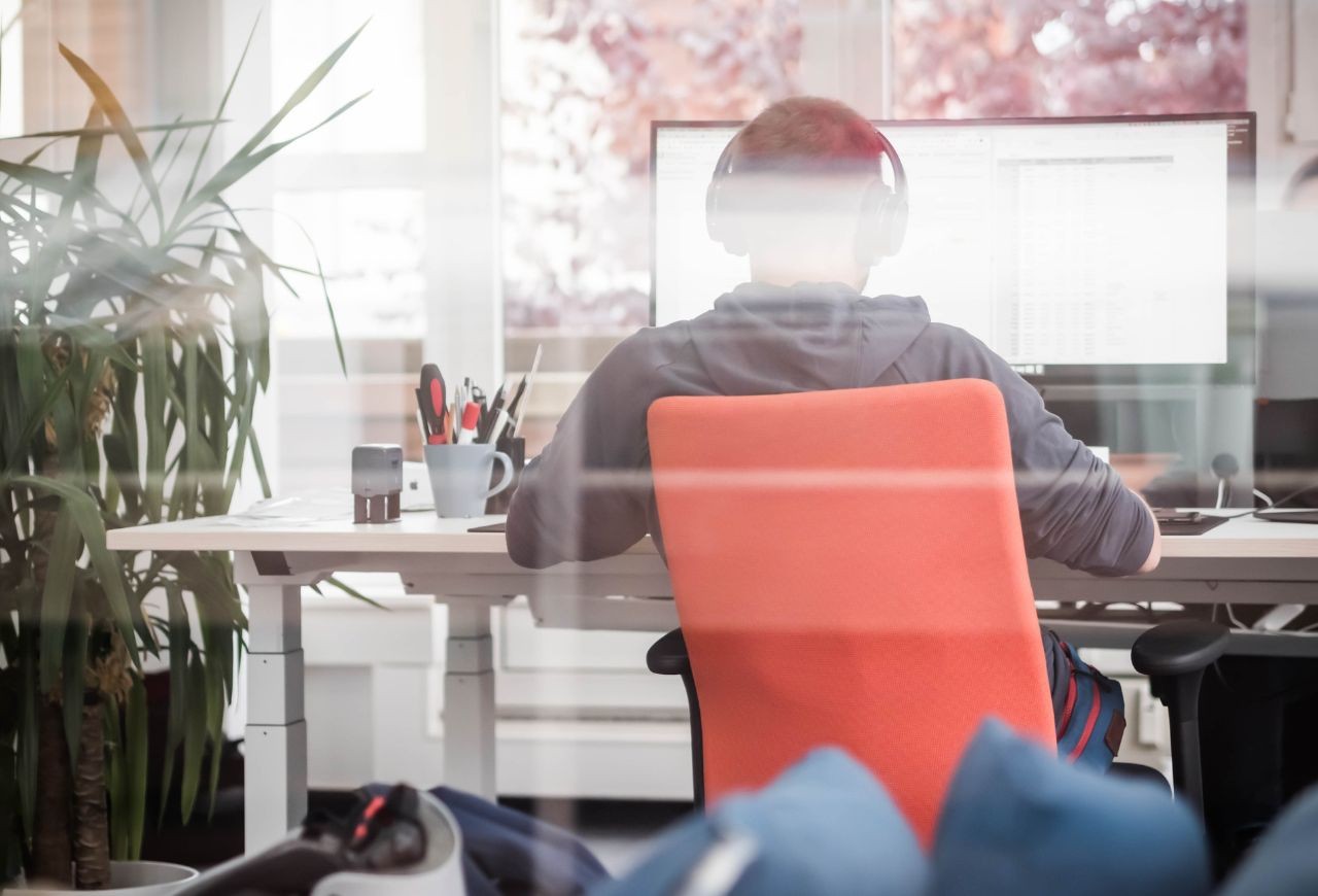 IT-Servicemitarbeiter mit Kopfhörern sitzt an einem Schreibtisch mit einem großen Bildschirm, Pflanzen und Büromaterial im Hintergrund.