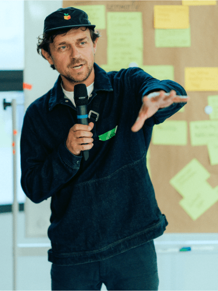 MANDARIN-Geschäftsleiter hält eine Präsentation mit Mikrofon, gestikulierend vor einer Pinnwand mit bunten Sticky Notes während eines Workshops.