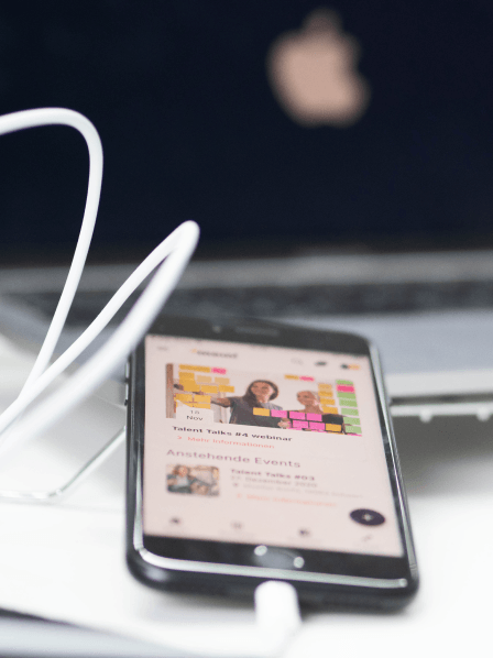 Smartphone mit geöffneter Event-Seite, die einen Webinar-Workshop zeigt, im Vordergrund eines Arbeitsplatzes mit Laptop und Ladekabel.