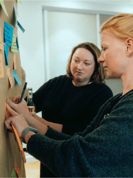 Zwei MANDARIN-Mitarbeiterinnen bei einem kreativen Workshop, arbeiten konzentriert an einer Pinnwand mit bunten Sticky Notes in einem modernen Meetingraum.