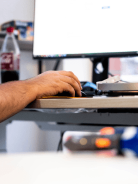 Hand eines Mitarbeiters am Schreibtisch, arbeitend mit Maus und Tastatur vor einem Bildschirm, im Hintergrund eine geöffnete Getränkeflasche und Bürozubehör.