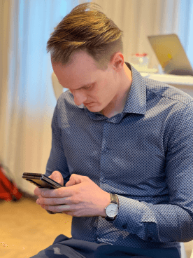 MANDARIN-Mitarbeiter konzentriert am Smartphone, in einem modernen Büroambiente mit Laptop und Arbeitsmaterialien im Hintergrund.