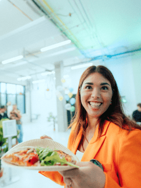 Fröhliche MANDARIN-Kollegin bei einem Event, hält einen Teller mit Pizza und Salat, in einem modernen Veranstaltungsraum mit dekorativen Lichtern und Ballons im Hintergrund.