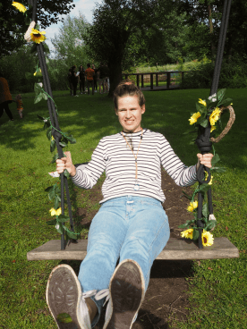 MANDARIN-Mitarbeiter*in entspannt auf einer mit Blumen dekorierten Schaukel während eines Outdoor-Events, umgeben von grüner Natur und sonnigem Wetter.