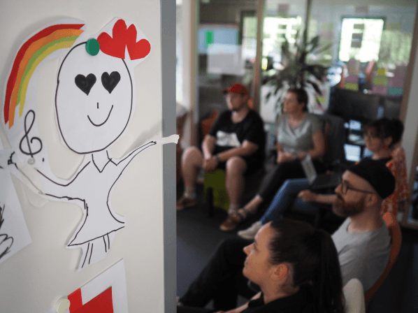 Kreativer Workshop bei MANDARIN, Fokus auf eine bunte Zeichnung mit Regenbogen und Herz, im Hintergrund Mitarbeiter*innen in einer Brainstorming-Session in einem modernen Büro.