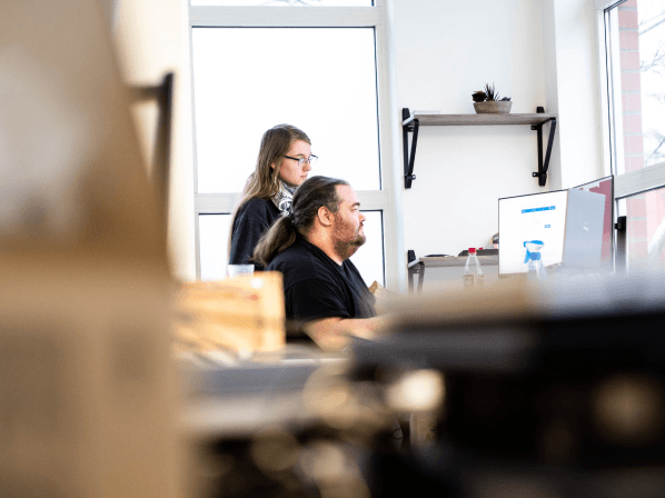 MANDARIN-Mitarbeiter*innen von der IT bei der gemeinsamen Arbeit am Computer, in einem modernen Büro mit großen Fenstern, Regalen und heller Atmosphäre.