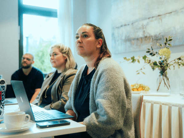 MANDARIN-Mitarbeiter*innen bei einem Meeting, aufmerksam einem Vortrag folgend, mit Laptops und Notizen an einem modernen Arbeitsplatz, dekoriert mit Blumen und natürlichem Tageslicht.