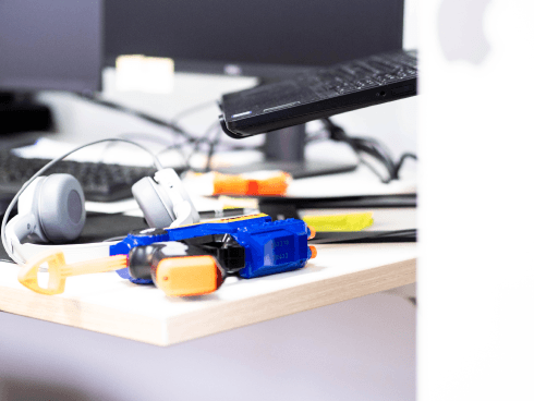 Schreibtisch im MANDARIN-Büro mit Kopfhörern, Laptop, Monitoren und einer Spielzeug-Nerf-Gun, kombiniert mit Bürozubehör für eine kreative Arbeitsatmosphäre.