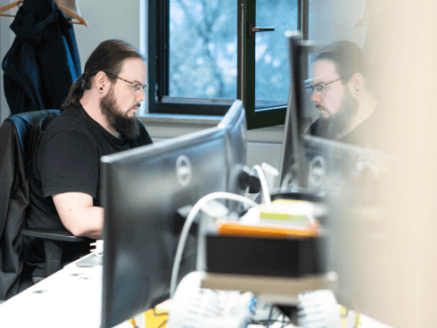 MANDARIN-Mitarbeiter konzentriert am Arbeitsplatz mit zwei Monitoren, in einem modernen Büro mit Blick nach draußen durch ein geöffnetes Fenster.