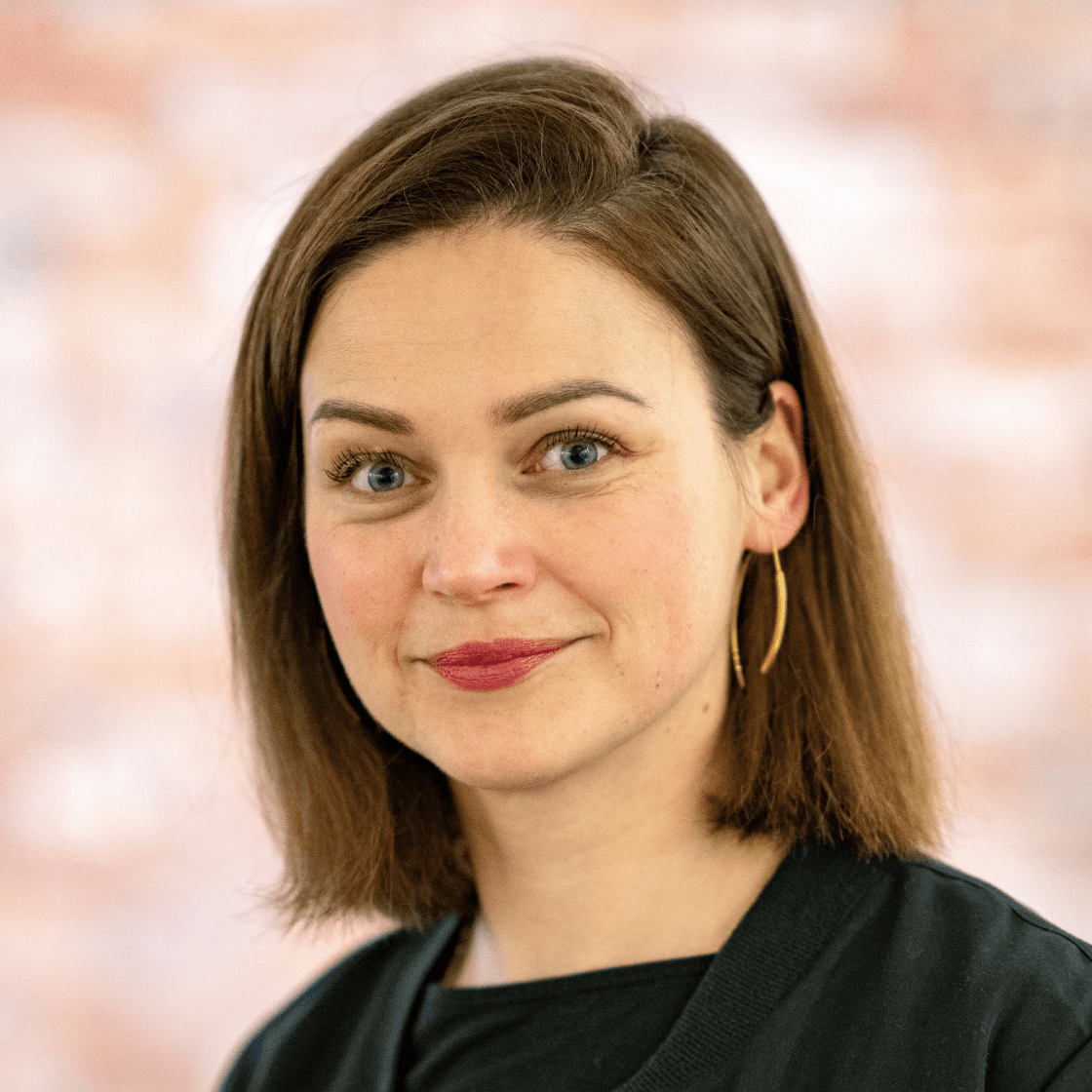 Dieses Porträt zeigt Anne, eine junge Frau mit schulterlangem braunem Haar und grünen Augen. Sie trägt ein schwarzes Oberteil und goldene Ohrringe, die ihr Gesicht elegant betonen. Ihr freundlicher Gesichtsausdruck und das dezente Lächeln verleihen dem Bild eine sympathische und authentische Ausstrahlung. Der unscharfe, warme Hintergrund sorgt für eine angenehme Bildtiefe und hebt Anne als Hauptmotiv hervor. 