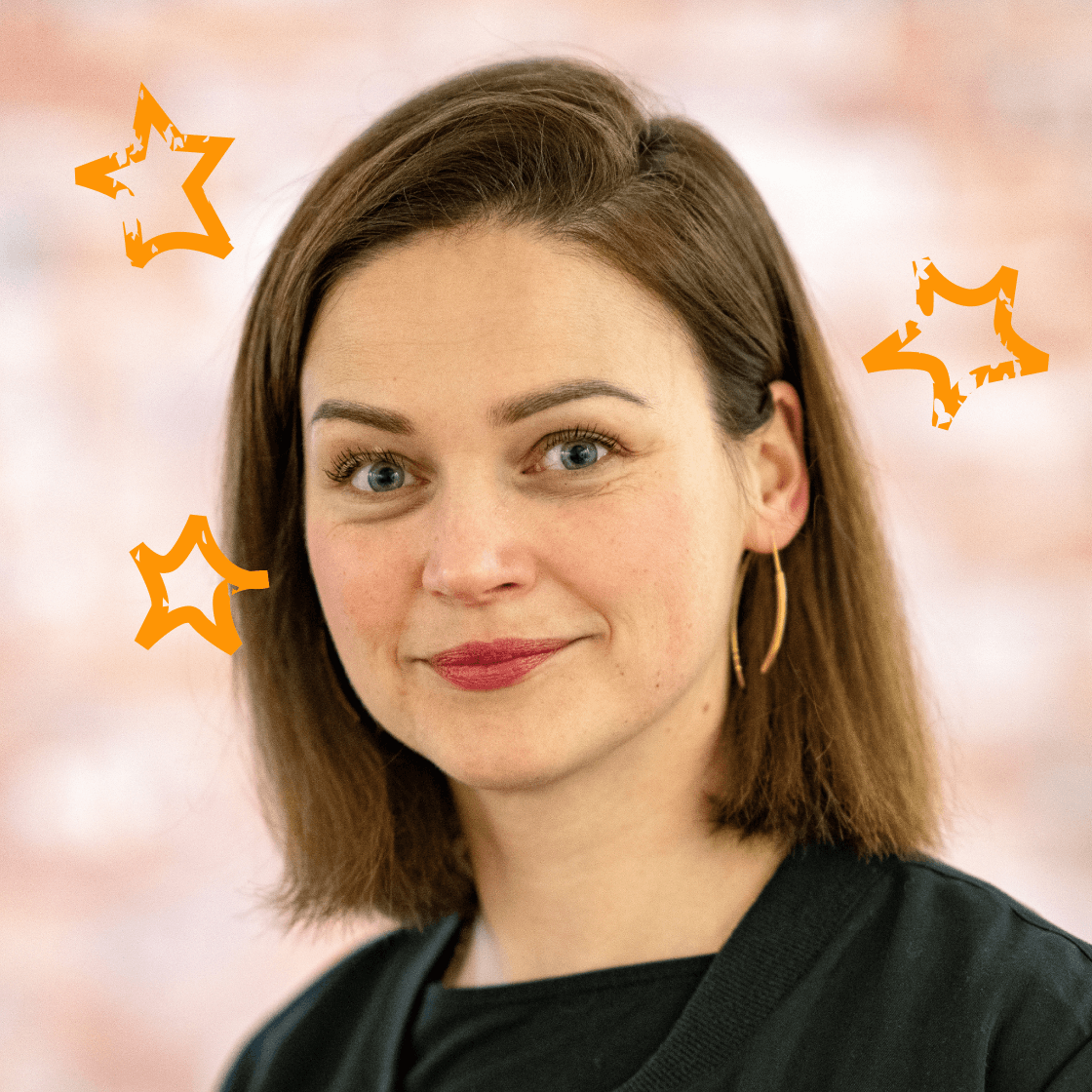 Dieses Porträt zeigt Anne, eine junge Frau mit schulterlangem braunem Haar und grünen Augen. Sie trägt ein schwarzes Oberteil und goldene Ohrringe, die ihr Gesicht elegant betonen. Ihr freundliches Lächeln verleiht dem Bild eine sympathische und authentische Ausstrahlung. Der warme, unscharfe Hintergrund sorgt für eine angenehme Bildtiefe, während die hinzugefügten orangefarbenen Sterne einen kreativen, dynamischen Effekt erzeugen.