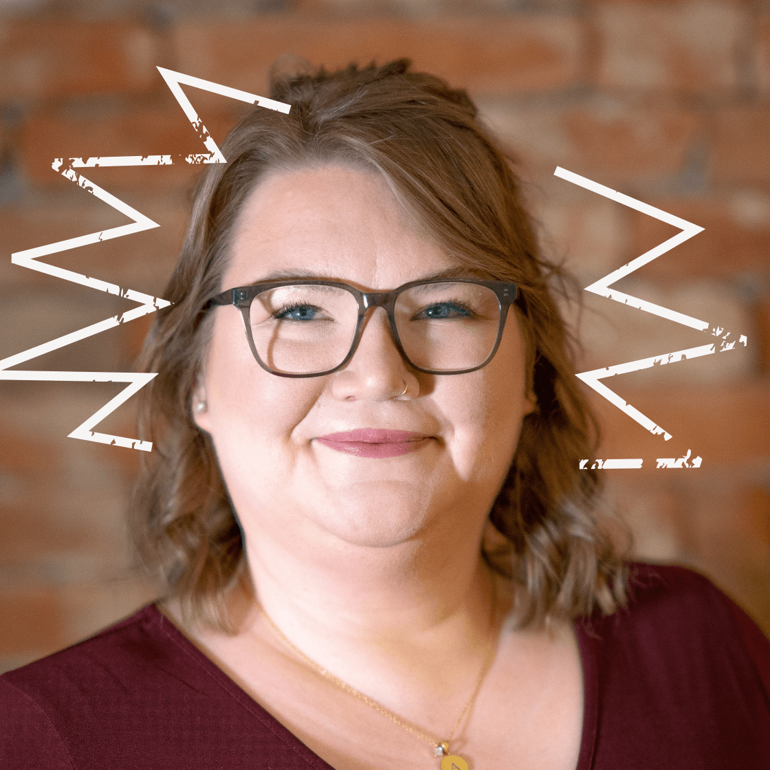 Dieses Porträt zeigt Annika, eine Frau mit schulterlangem, welligem Haar, Brille und einem warmen Lächeln. Sie trägt ein weinrotes Oberteil und eine goldene Kette mit Anhänger, die ihrem Look eine elegante und persönliche Note verleiht. Der warme Backstein-Hintergrund sorgt für eine moderne, authentische Atmosphäre. Ein kreatives Detail: Die gezeichneten dynamischen Linien um ihren Kopf verleihen dem Bild eine energiegeladene, ausdrucksstarke Wirkung.