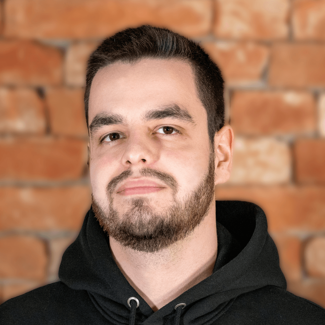 Dieses Porträt zeigt Dennis, einen jungen Mann mit dunklem Haar, gepflegtem Bart und freundlichem Ausdruck. Er trägt einen schwarzen Hoodie, der einen lässigen und modernen Look verleiht. Der warme Backstein-Hintergrund sorgt für eine rustikale und zugleich professionelle Atmosphäre. Die weiche Beleuchtung hebt sein Gesicht vorteilhaft hervor und verleiht dem Bild eine authentische, selbstbewusste Ausstrahlung.