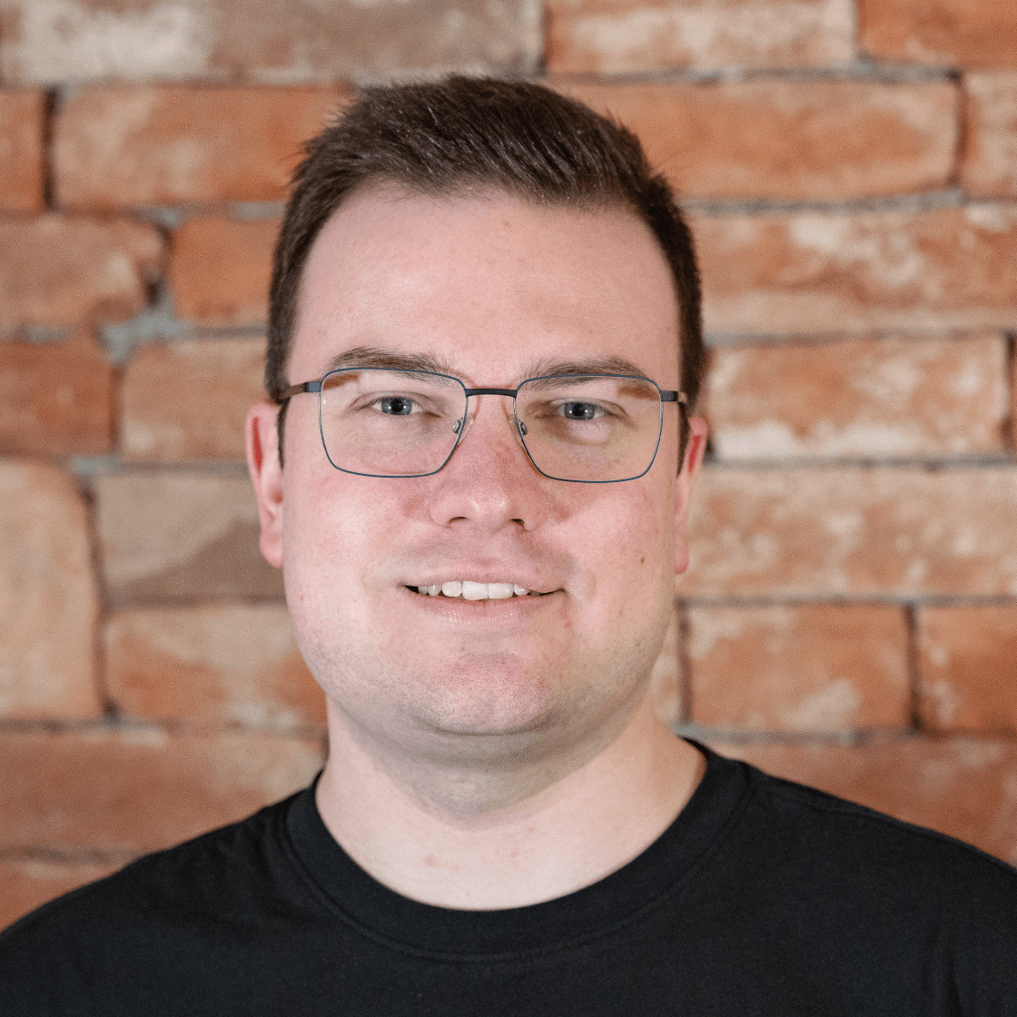 Dieses Porträt zeigt Hannes, einen Mann mit kurzem, dunkelblondem Haar, Brille und einem freundlichen Lächeln. Er trägt ein schwarzes Oberteil, das für einen modernen und professionellen Look sorgt. Der warme Backstein-Hintergrund schafft eine authentische, stilvolle Atmosphäre und hebt Hannes als Hauptmotiv hervor. Die natürliche Beleuchtung betont seine Gesichtszüge und verleiht dem Bild eine sympathische und selbstbewusste Ausstrahlung.