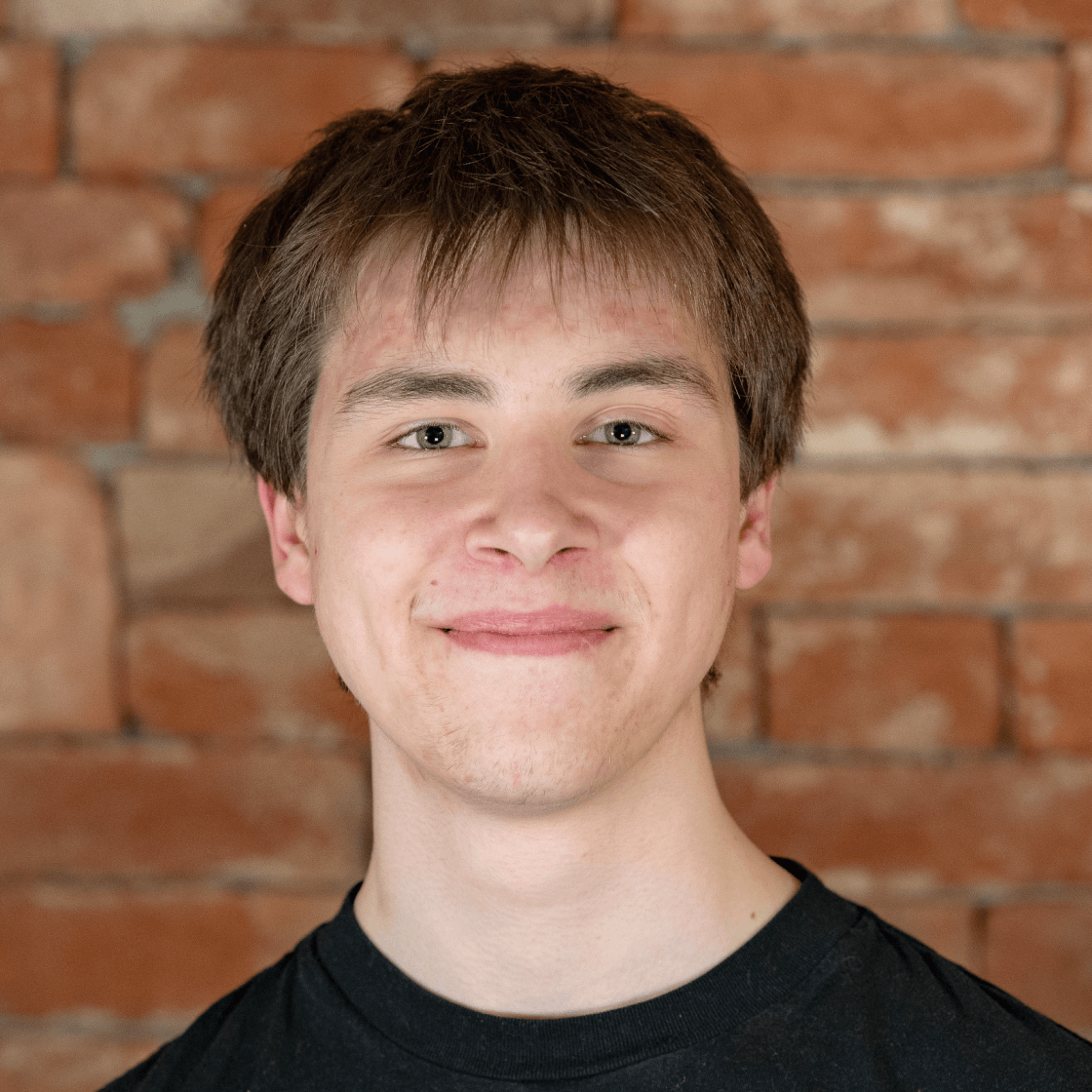 Dieses Porträt zeigt Steven, einen jungen Mann mit mittellangem braunem Haar und einem freundlichen Lächeln. Er trägt ein schwarzes T-Shirt, das einen lässigen und modernen Look vermittelt. Der warme Backstein-Hintergrund schafft eine authentische, einladende Atmosphäre und hebt Steven als Hauptmotiv hervor. Die natürliche Beleuchtung betont seine Gesichtszüge und sorgt für eine sympathische, authentische Ausstrahlung.