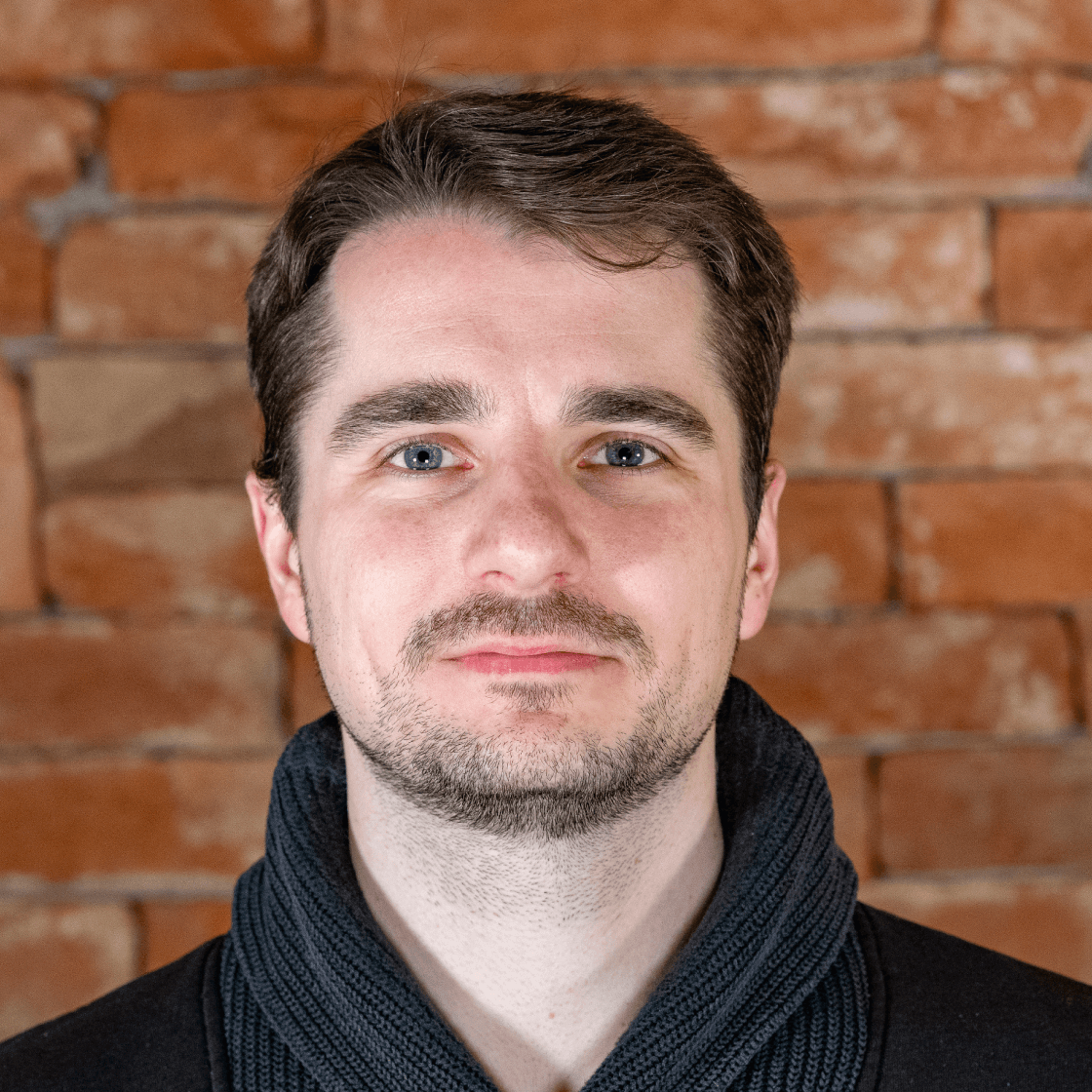 Dieses Porträt zeigt Veit, einen Mann mit kurzen, braunen Haaren, dezentem Bart und blauen Augen. Er trägt einen dunklen Pullover mit Schalkragen, der einen stilvollen und professionellen Look verleiht. Der warme Backstein-Hintergrund schafft eine authentische, moderne Atmosphäre und hebt Veit als Hauptmotiv hervor. Die sanfte Beleuchtung betont seine Gesichtszüge und sorgt für eine natürliche, seriöse Ausstrahlung. 
