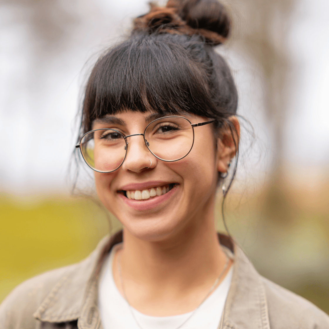 Porträt von Christina mit dunklem, hochgestecktem Haar und geradem Pony. Sie trägt eine runde Brille, einen Nasenring sowie Ohrringe und lächelt freundlich in die Kamera. Ihr Outfit besteht aus einem hellen Shirt und einer beigefarbenen Jacke. Der Hintergrund ist unscharf und zeigt eine grüne Naturkulisse.