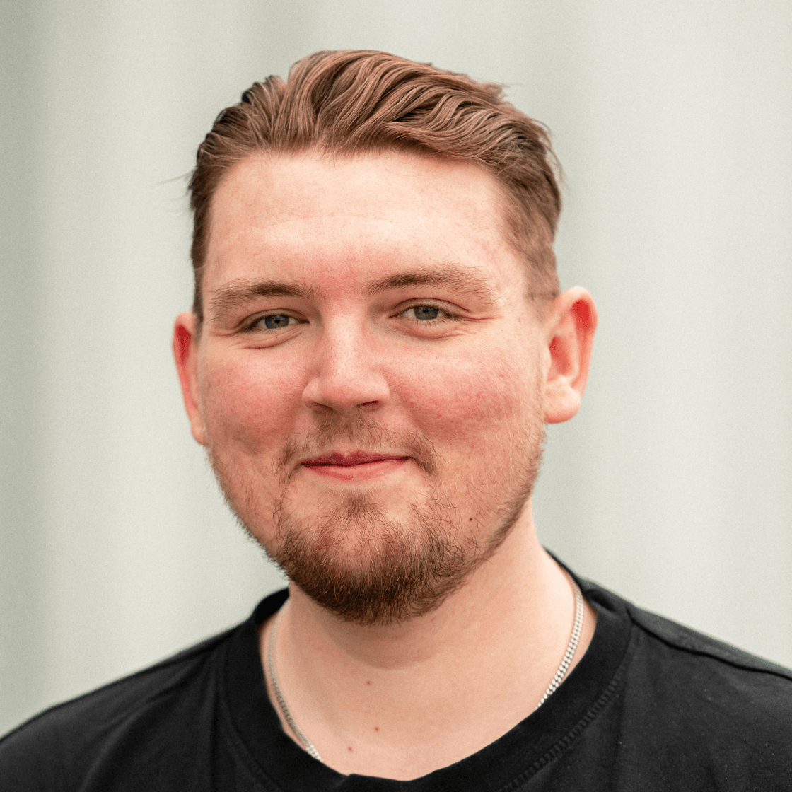 Porträt von Friedrich mit kurz geschnittenem, zurückgestyltem Haar und gepflegtem Bart. Er trägt ein schwarzes T-Shirt und eine silberne Kette. Sein Blick ist freundlich, der Hintergrund ist neutral und leicht verschwommen.