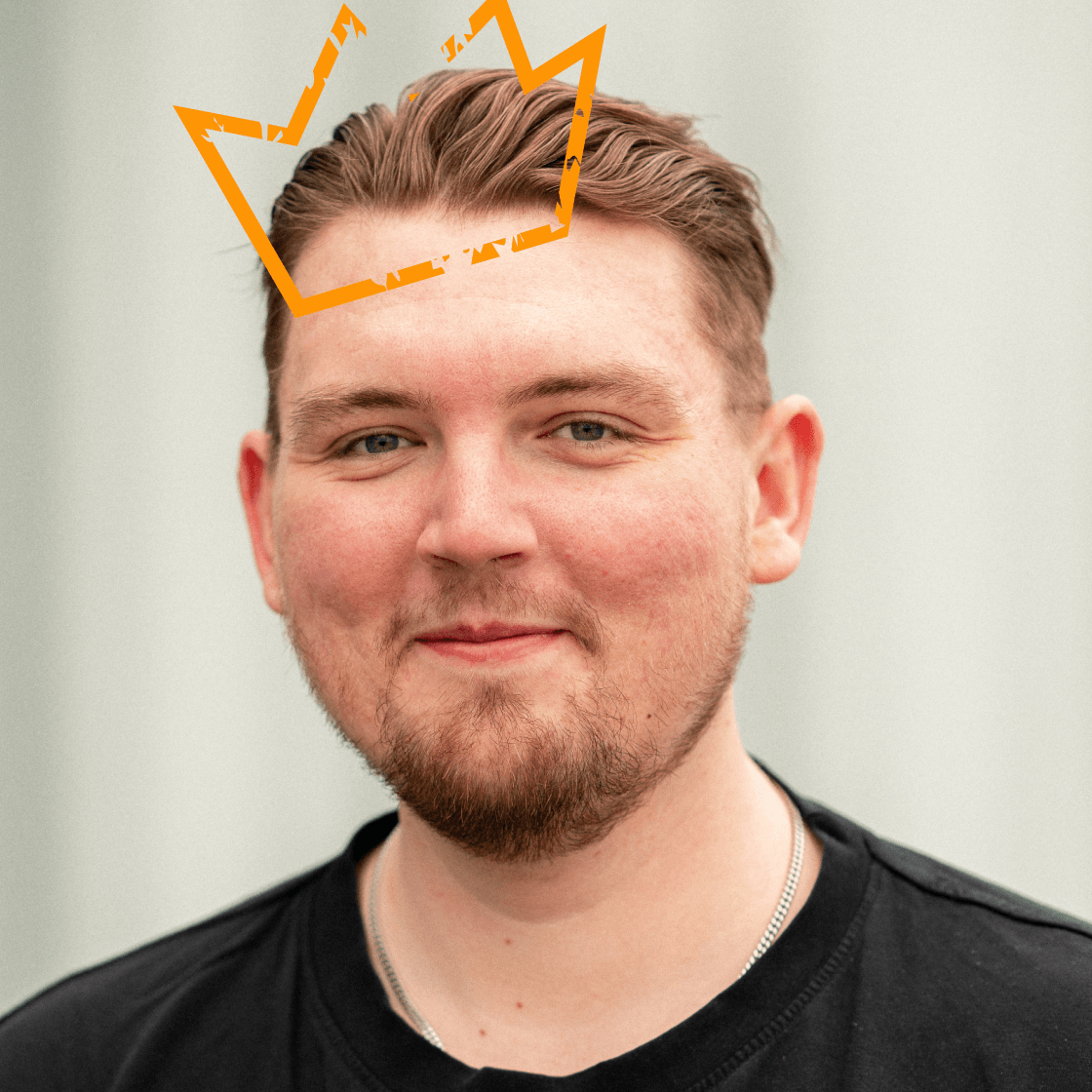 Porträt von Friedrich mit zurückgestyltem Haar, gepflegtem Bart und schwarzem T-Shirt. Über seinem Kopf schwebt eine stilisierte, orangefarbene Krone – Symbol für Kompetenz oder Auszeichnung. Freundlicher Gesichtsausdruck vor hellem, neutralem Hintergrund.