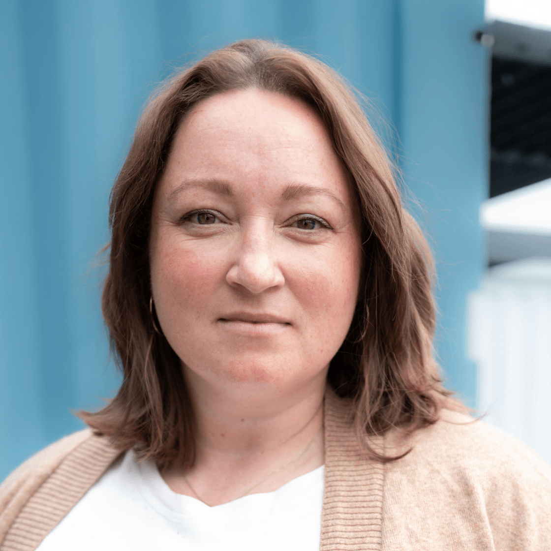 Porträt von Julia mit mittellangem braunem Haar, hellem Shirt und beigefarbener Strickjacke. Sie blickt ruhig und selbstbewusst in die Kamera. Der Hintergrund zeigt eine blaue Containerwand, die für eine moderne, klare Atmosphäre sorgt.