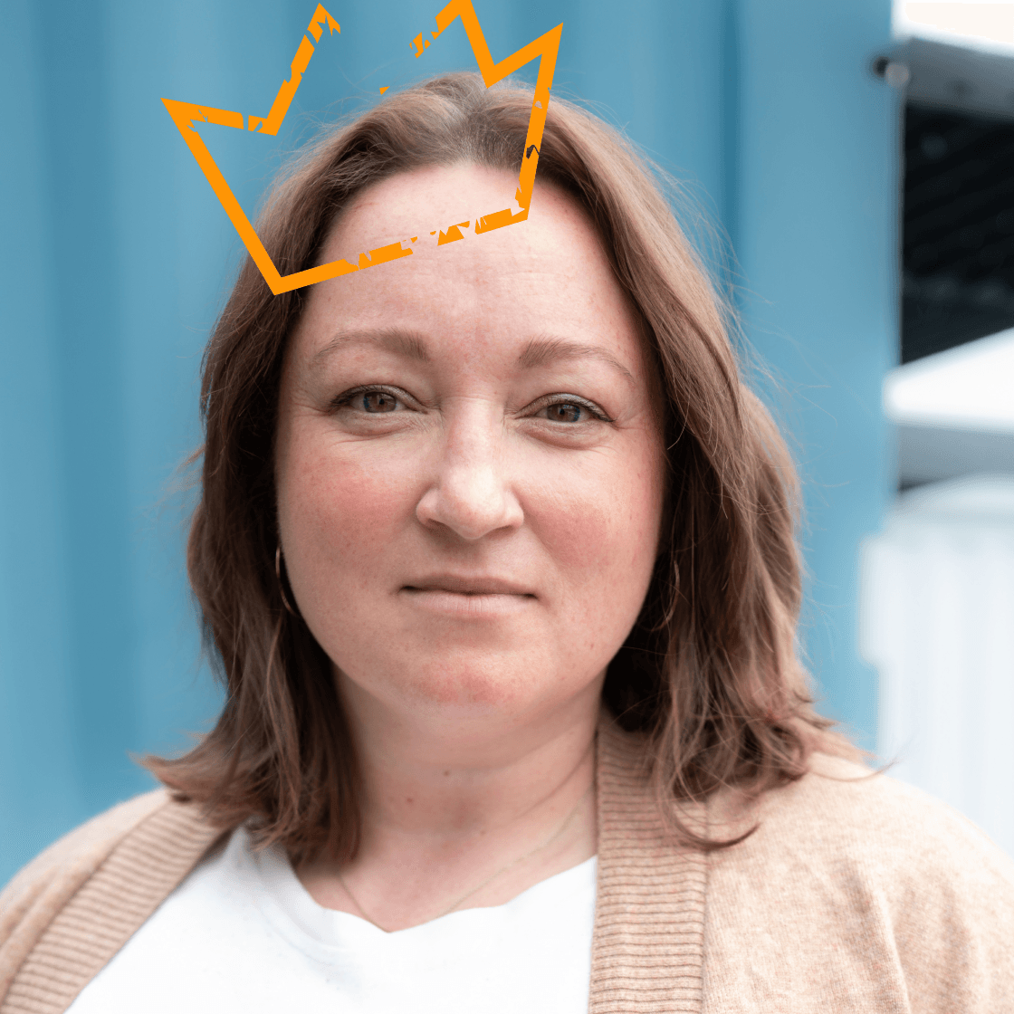 Porträt von Julia mit schulterlangem braunem Haar, beiger Strickjacke und weißem Shirt. Über ihrem Kopf ist eine stilisierte, orangefarbene Krone platziert – ein Symbol für Kompetenz, Leadership oder besondere Wertschätzung. Im Hintergrund ist eine moderne, hellblaue Containerwand zu sehen.