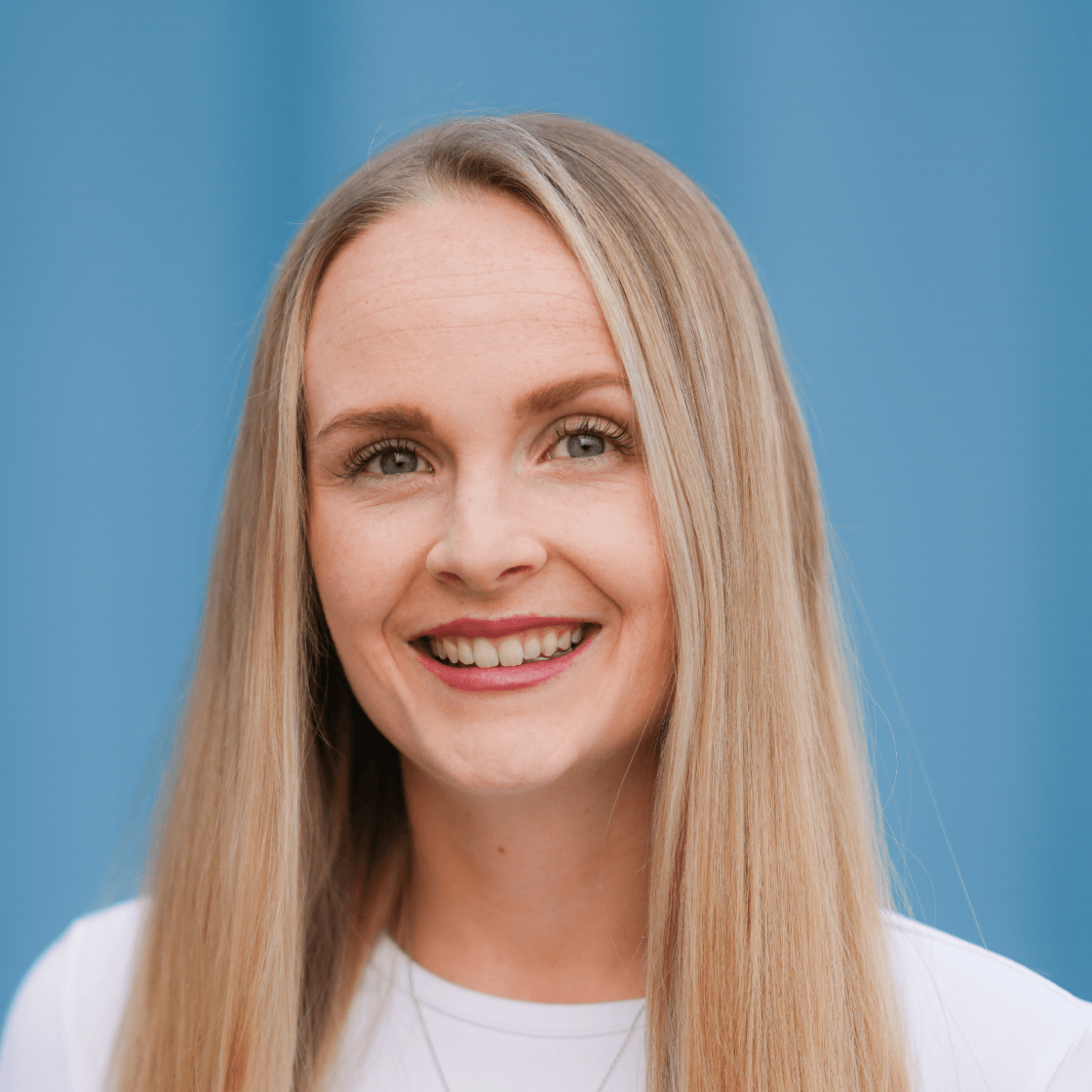  „Porträt von Juliana mit langen, glatten, blonden Haaren und einem freundlichen Lächeln. Sie trägt ein weißes Oberteil und eine dezente Halskette. Der Hintergrund ist einfarbig in einem klaren Blauton gehalten und sorgt für einen modernen, frischen Look.