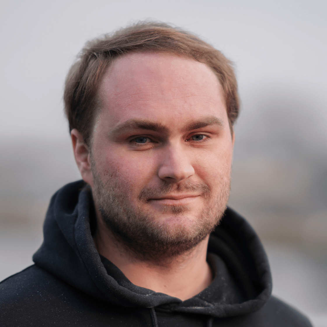 Porträt von Max mit kurzem, hellbraunem Haar und dunklem Hoodie. Er blickt ruhig und selbstbewusst in die Kamera. Der Hintergrund ist dezent verschwommen und zeigt eine neblig-graue Außenkulisse, die dem Bild eine ruhige, authentische Atmosphäre verleiht