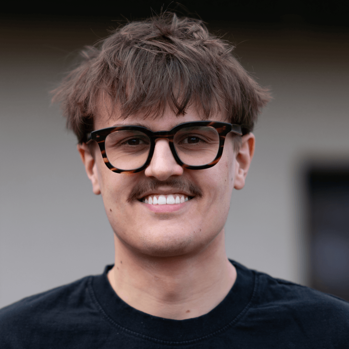  „Porträt von Toni mit braunen Haaren, markanter Brille im Havanna-Stil und einem dezenten Schnurrbart. Toni trägt ein schwarzes T-Shirt und lächelt sympathisch in die Kamera. Der Hintergrund ist neutral und weichgezeichnet – ideal für einen professionellen, aber nahbaren Eindruck