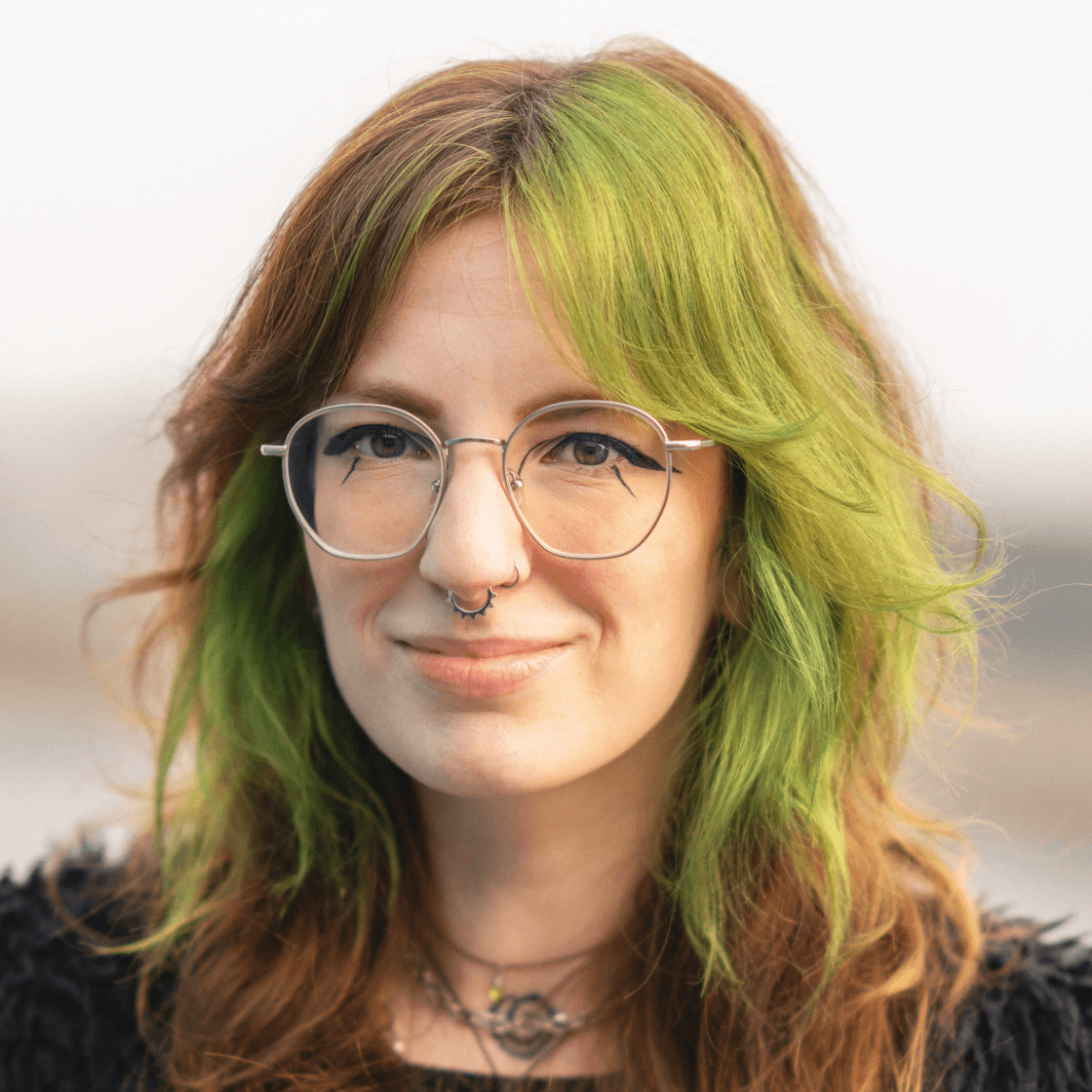 Porträt von Valeria mit grün-braunem Haar, runder Brille und Septum-Piercing. Sie trägt dunkle Kleidung und eine Kette mit Anhängern. Ihr Gesichtsausdruck ist freundlich und selbstbewusst, im Hintergrund ist ein unscharfer, heller Outdoor-Hintergrund zu sehen.