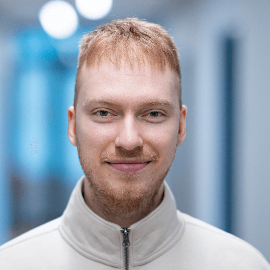 Porträt von Niklas mit blonden, kurzen Haaren, Bart und hellem Zipper-Pullover. Er lächelt ruhig und freundlich in die Kamera. Der Hintergrund zeigt einen modernen, unscharfen Büroflur mit kühler Lichtstimmung.