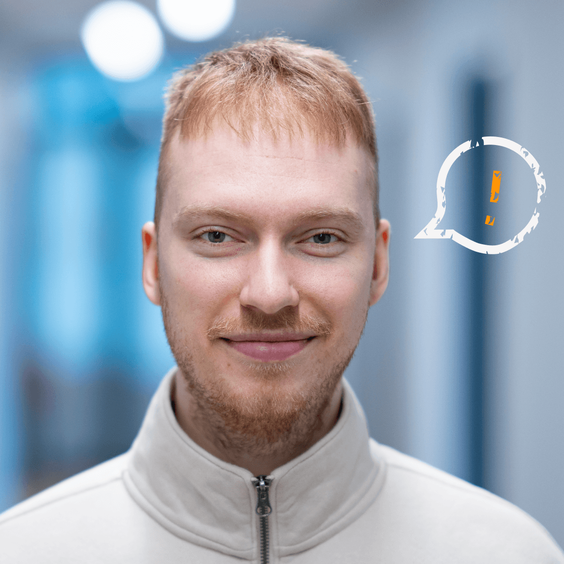 „Porträt von Niklas mit kurzen blonden Haaren, Bart und hellem Zipper-Pullover. Er lächelt entspannt in die Kamera. Rechts neben ihm ist eine stilisierte weiße Sprechblase mit orangefarbenem Ausrufezeichen zu sehen – ein Symbol für wichtige Hinweise, Impulse oder Kommunikation. Der Hintergrund zeigt einen modernen Büroflur in kühler Lichtstimmung.